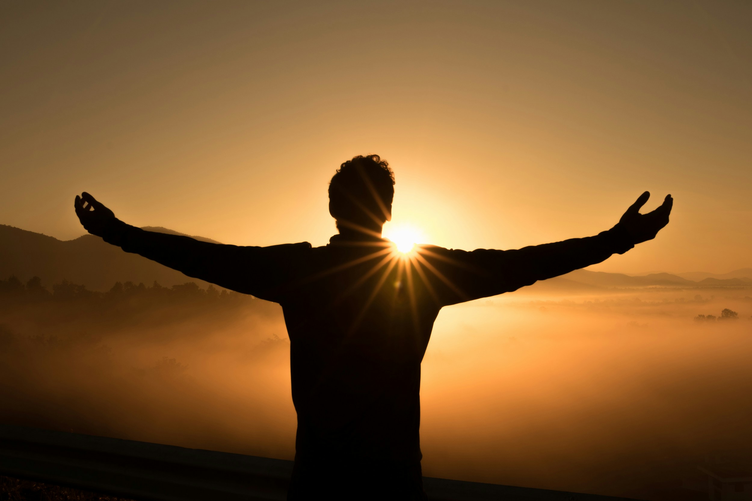 A person standing at sunrise with arms outstretched, overlooking a misty valley