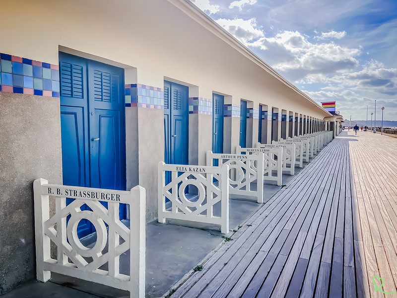 Golden hour on the Deauville boardwalk in Normandy, France, with its iconic red-and-white striped cabanas lining the pebble beach and the English Channel stretching to the horizon