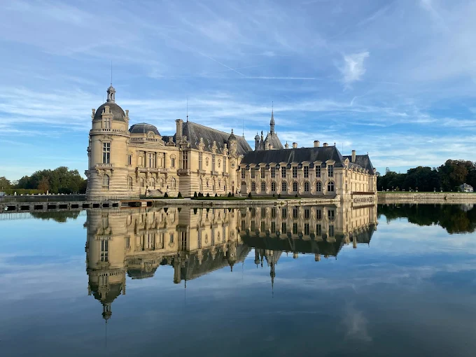 Chantilly Castle at golden hour in France, its grand pink-brick façade reflected in the still waters of the moat with manicured gardens stretching into the distance