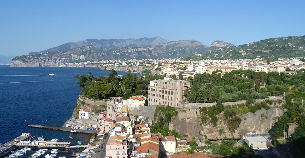 Dramatic Sorrento cliffs plunging into the turquoise Tyrrhenian Sea, with colorful buildings perched on the edge and a small beach below at mid-morning