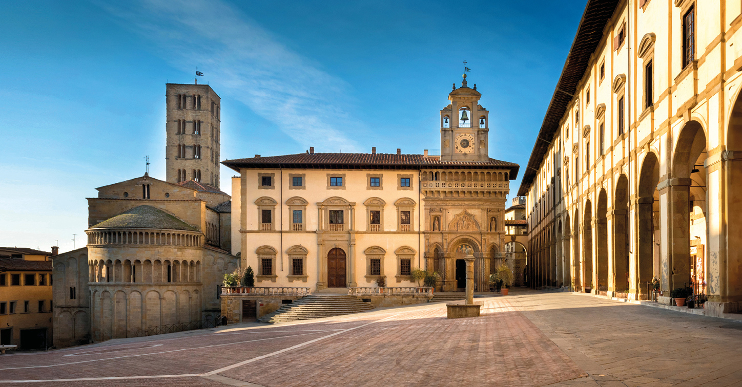 Sun-drenched Piazza Grande in Arezzo, Italy, showcasing medieval buildings, the Loggia Vasari, and sloping cobblestones at mid-morning