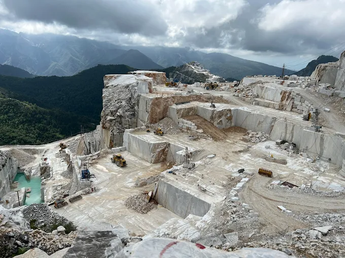 A vast, sunlit Carrara marble quarry in the Apuan Alps, with stark white rock faces and a small red vehicle for scale, mid-morning