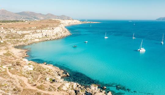 Turquoise waters of Cala Azzurra beach on Favignana island, with dramatic limestone cliffs and a small sandy cove under a clear mid-morning sky