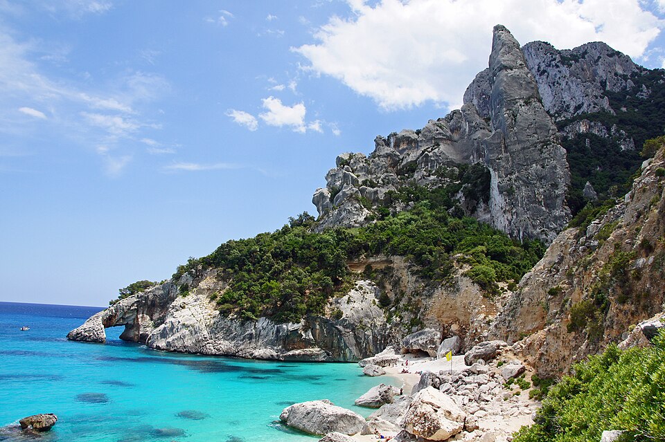 Dramatic Cala Goloritzé beach with turquoise water, white pebbles, and a towering limestone pinnacle under a clear mid-morning sky in Sardinia
