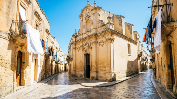 Sun-drenched labyrinth of Bari old town with narrow cobbled streets, laundry lines, and Baroque church facades under a clear blue sky at mid-morning