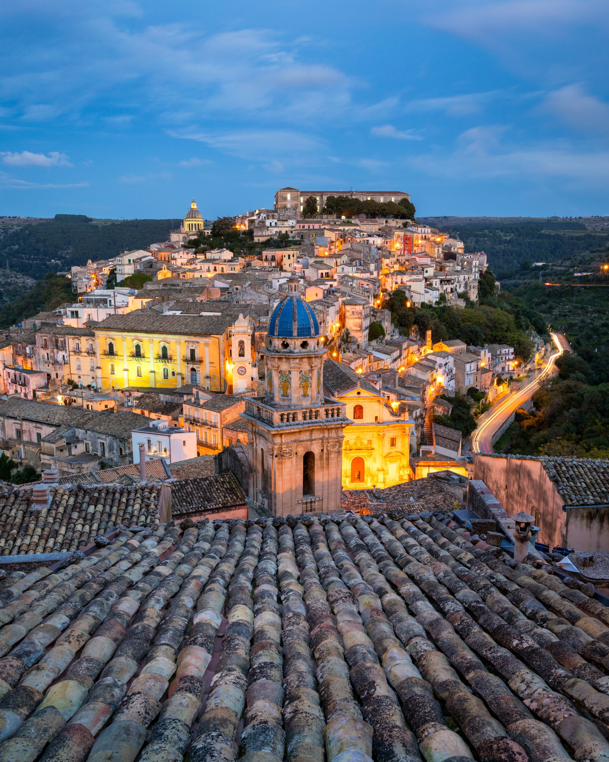 Sun-drenched Baroque buildings cascading down a hill in Ragusa Ibla, with terracotta roofs and the Hyblaean Mountains under a clear blue sky at mid-morning