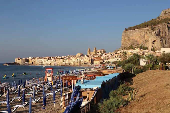 Cefalù beach with golden sand and turquoise water, backed by the medieval Duomo and La Rocca mountain under a clear mid-morning sky