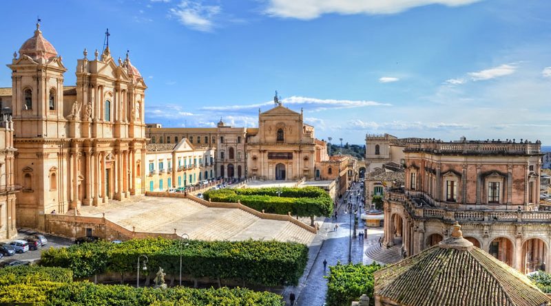 Sun-drenched Baroque buildings lining Corso Vittorio Emanuele in Noto, with golden limestone facades and ornate balconies under a clear blue sky at mid-morning