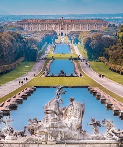 The grand Baroque facade of Caserta Royal Palace at mid-morning, with its symmetrical wings, fountains, and the long tree-lined avenue under a clear blue sky