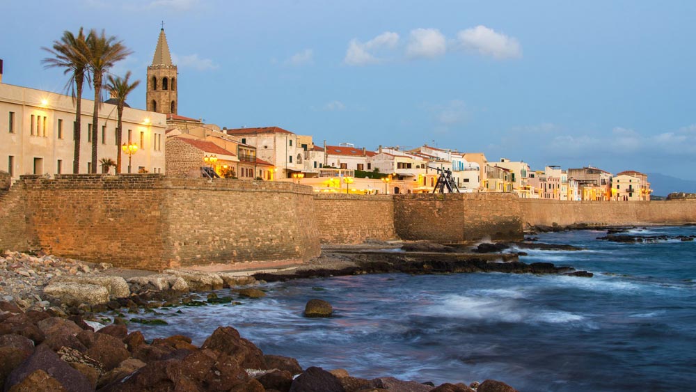 Sun-drenched Catalan quarter of Alghero with terracotta rooftops, narrow cobbled streets, and the Mediterranean Sea under a clear mid-morning sky in Sardinia