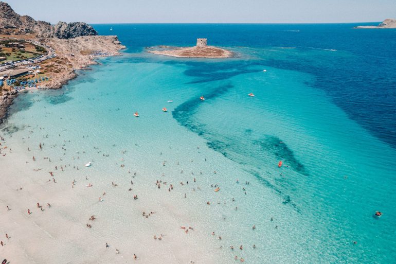 Turquoise waters and white sand of La Pelosa beach in Stintino, with dramatic sea stacks and clear Sardinian sky at mid-morning