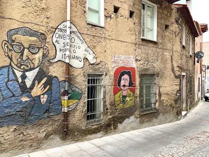 Morning light on Orgosolo murals in Sardinia's Barbagia region, vibrant political street art covering stone buildings under clear blue sky