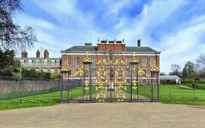 Kensington Palace red brick facade at golden hour with Kensington Gardens green lawn and London visitors walking