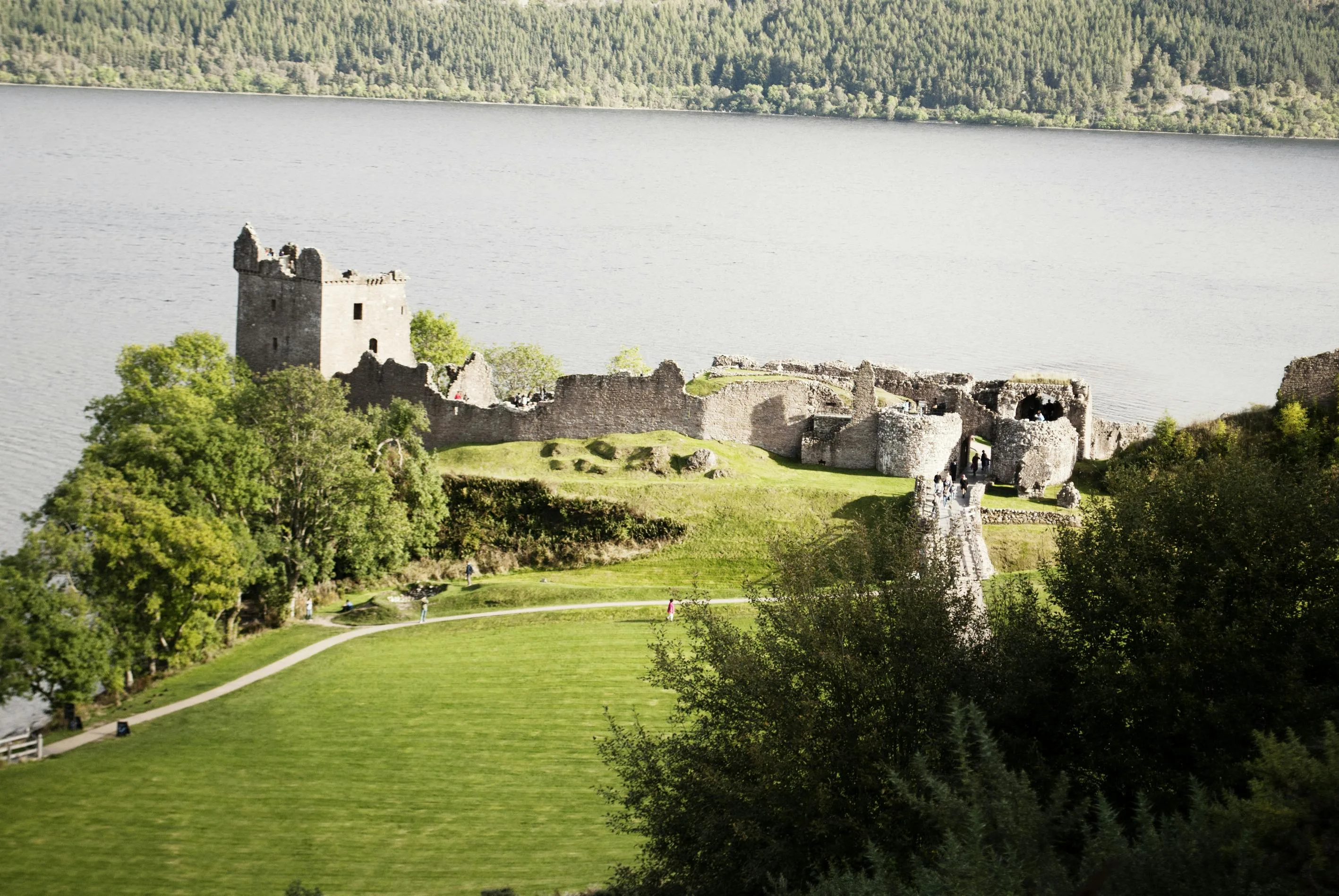 Urquhart Castle ruins at sunset overlooking Loch Ness with dark water, purple heather hills, and Scottish Highlands background