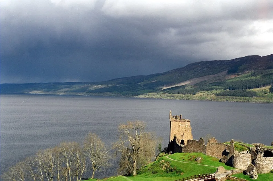 Misty Loch Ness waters at dawn with Urquhart Castle ruins, purple heather hills, and Scottish Highlands background