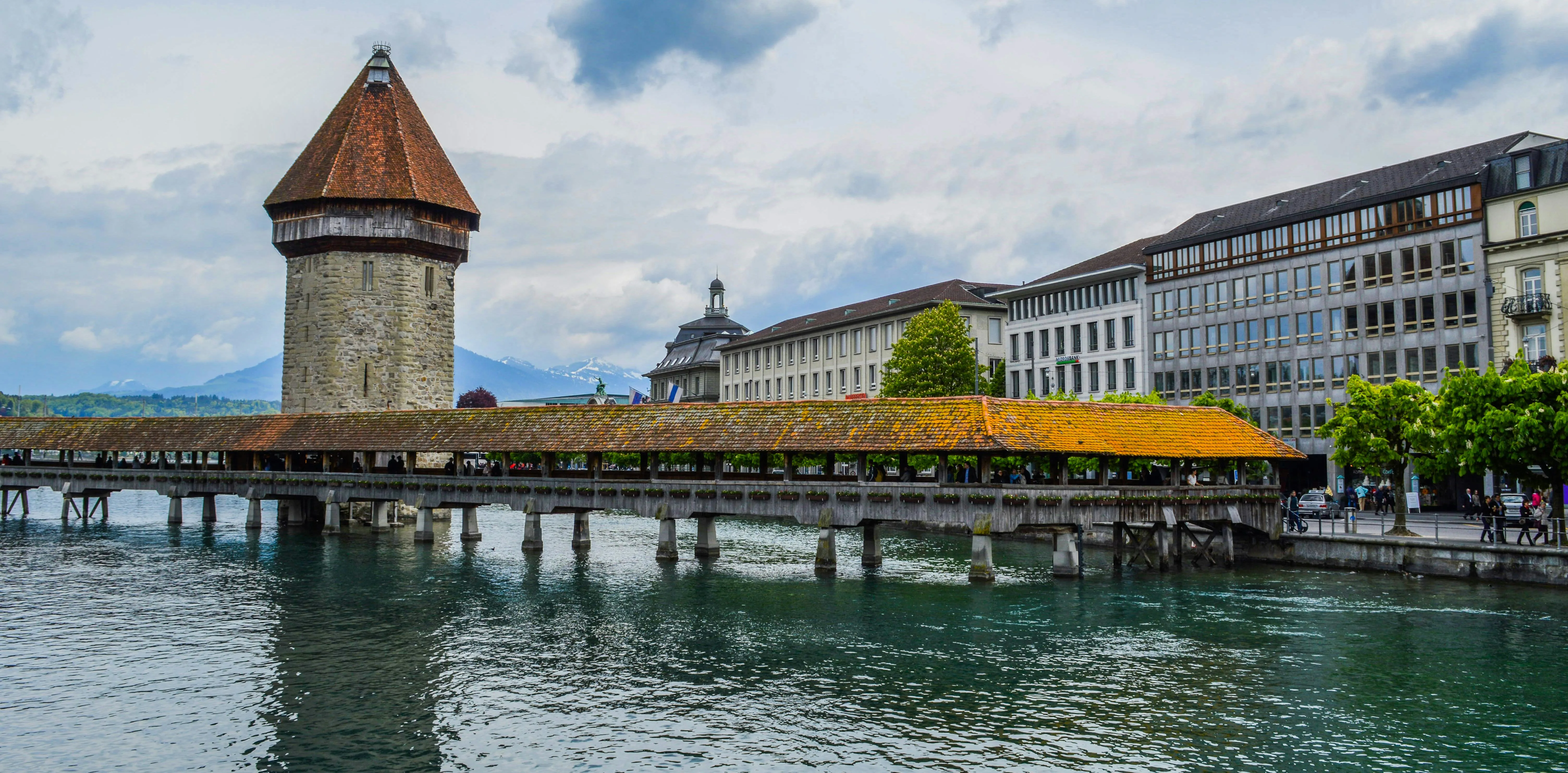 Lucerne Chapel Bridge at golden hour with octagonal Water Tower reflecting in Reuss River, historic triangular paintings visible under wooden roof, Swiss Alps backdrop