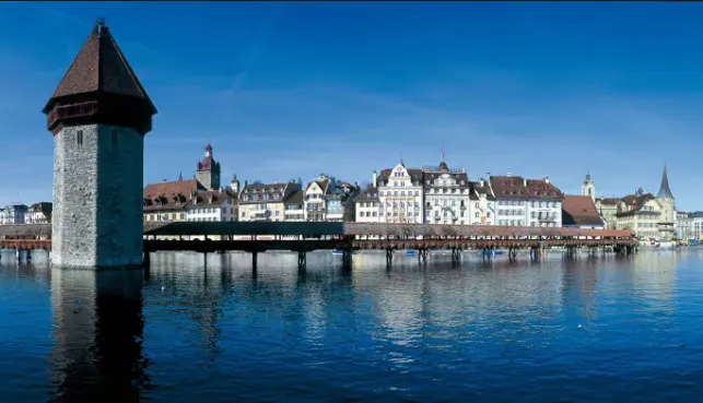 Lucerne Chapel Bridge wooden structure at sunrise with Reuss River, Wasserturm tower, and Alpine mountains in Switzerland