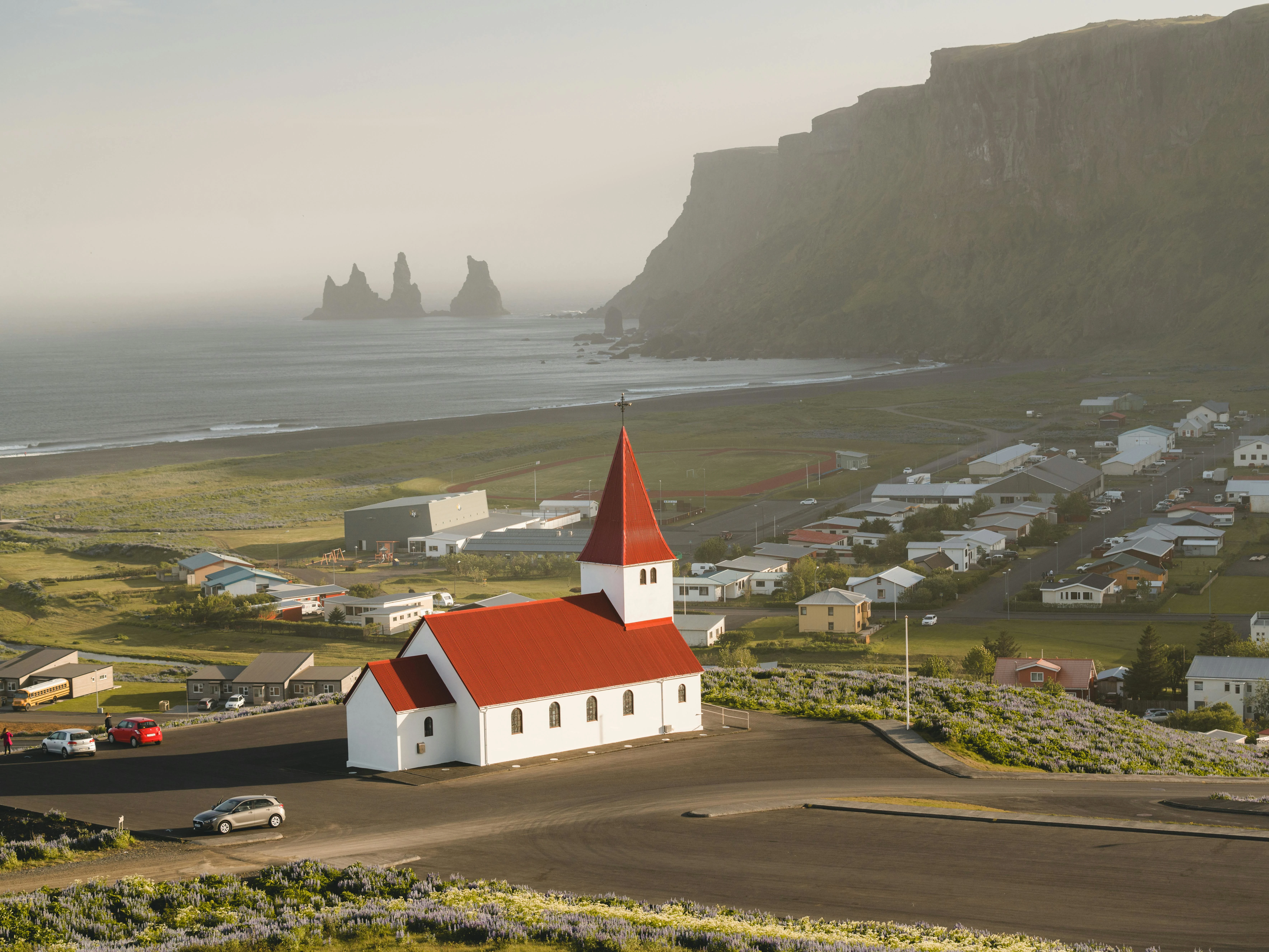 White Vik church with red roof on hill overlooking ocean and Reynisdrangar sea stacks under cloudy sky
