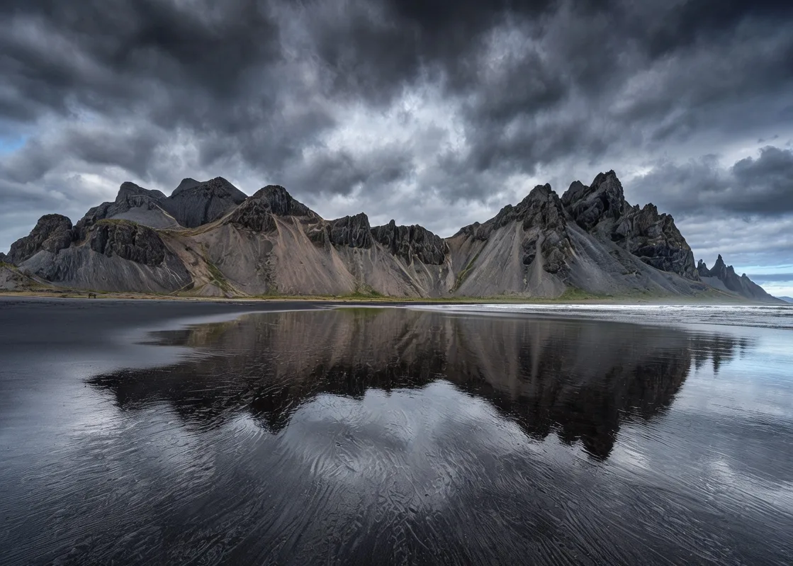 Vestrahorn mountain rising sharply from black sand beach with tidal reflections under moody Icelandic sky