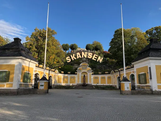 Skansen open-air museum historic wooden buildings surrounded by green trees at summer sunset with visitors walking